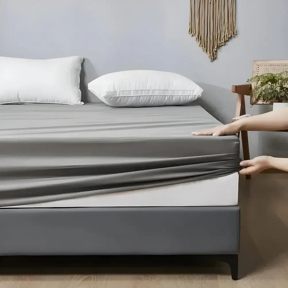 Imagem de uma cama com uma capa cinza sendo ajustada por duas mãos nos cantos do colchão. O colchão tem base cinza escura e dois travesseiros brancos no topo. A parede de fundo é cinza, decorada com uma peça de macramê e uma planta em uma cadeira ao lado.